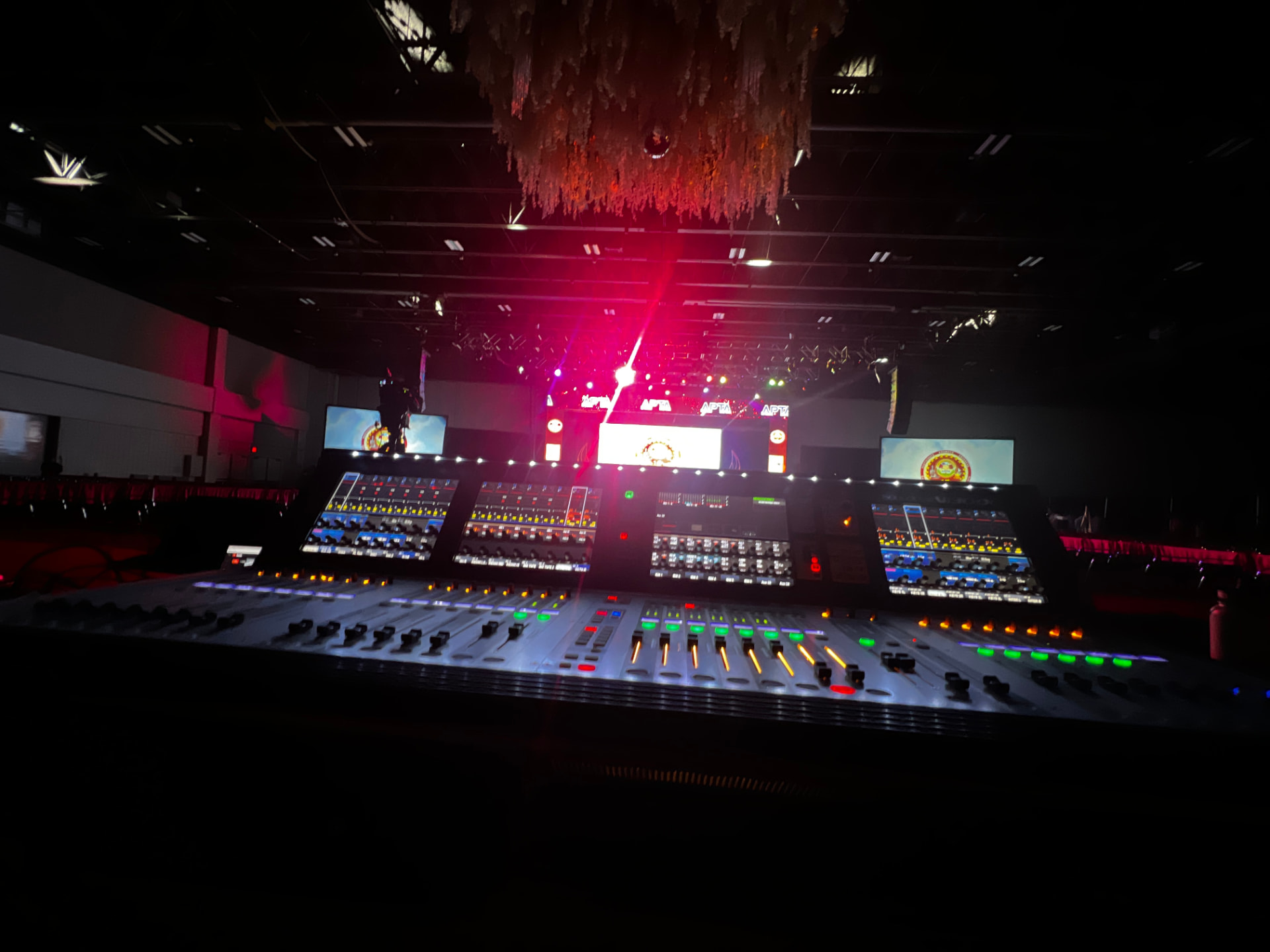 Two digital mixing consoles at front of house with a lit stage and LED screens in the background