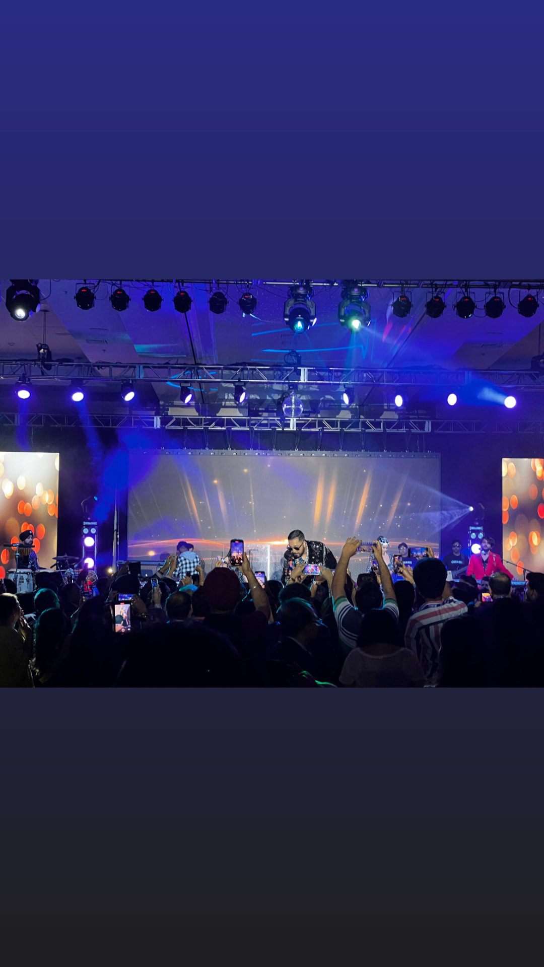 Indoor concert stage with blue moving-head lighting, LED wall and a packed crowd filming with phones